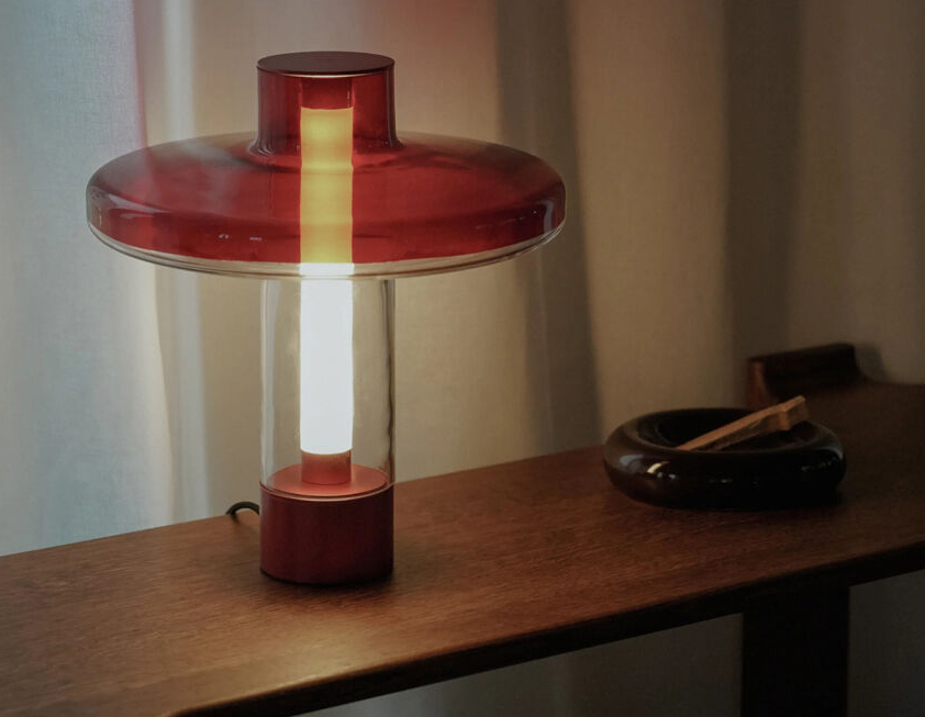 Nevo Table Lamp - Transparent Shade Creating a Stained Glass Effect 2 Nevo table lamp with red disc-shaped top and base, clear cylindrical body, and warm modern lighting on a wooden surface