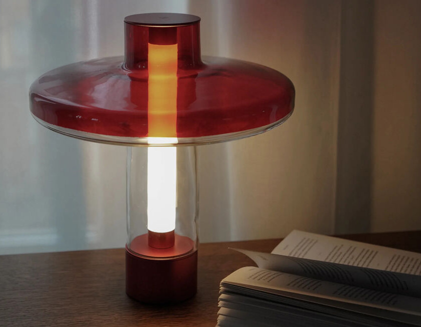 Nevo Table Lamp - Transparent Shade Creating a Stained Glass Effect 3 Nevo table lamp with red disc-shaped top and base, clear cylindrical body, and warm modern lighting on a wooden surface