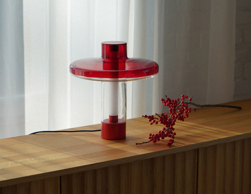 Nevo Table Lamp - Transparent Shade Creating a Stained Glass Effect 1 Nevo table lamp with red disc-shaped top and base, clear cylindrical body, and warm modern lighting on a wooden surface